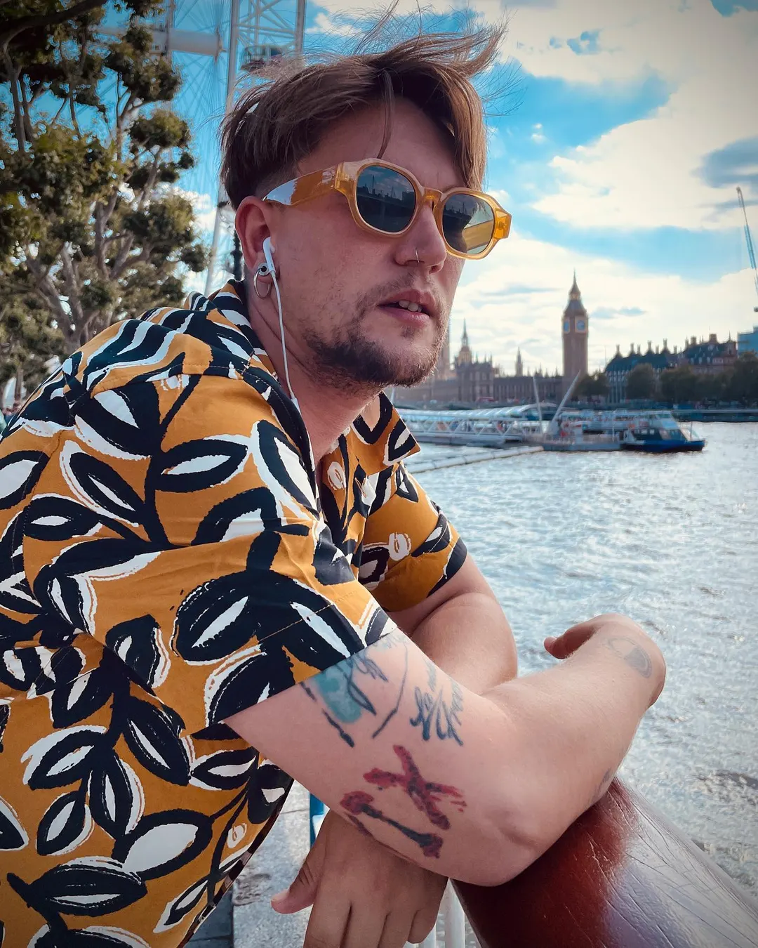 Cedecé apoyado en una baranda del Támesis con camisa estampada de hojas y gafas de sol amarillas, con el Big Ben al fondo.