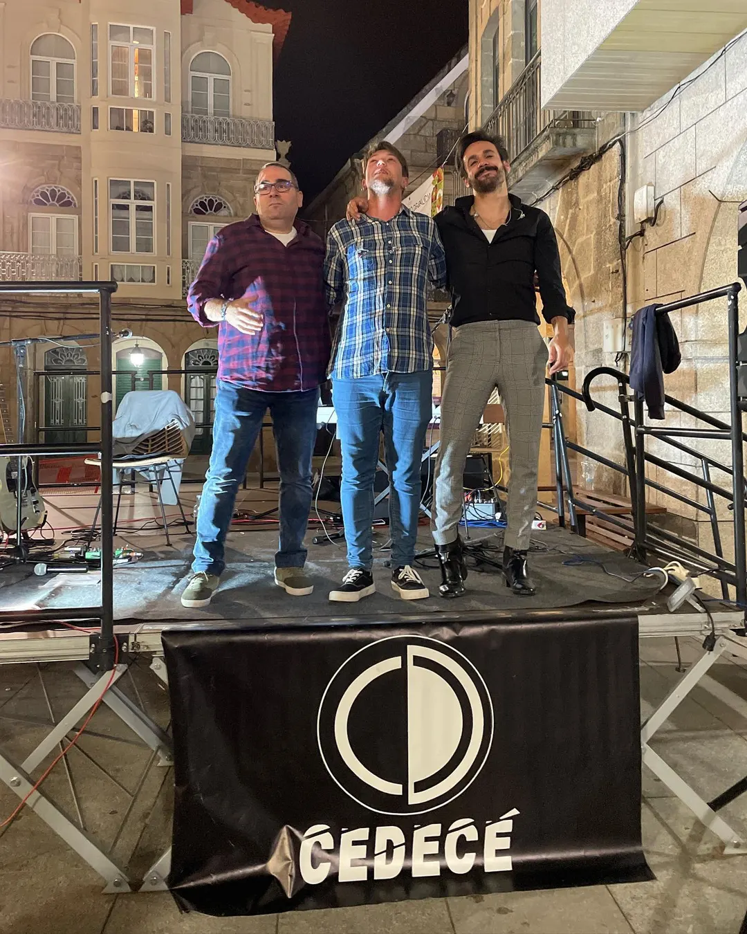 Los tres de pie sobre un escenario de noche en una plaza, tras un faldón negro con el logotipo circular y el rótulo CEDECÉ.