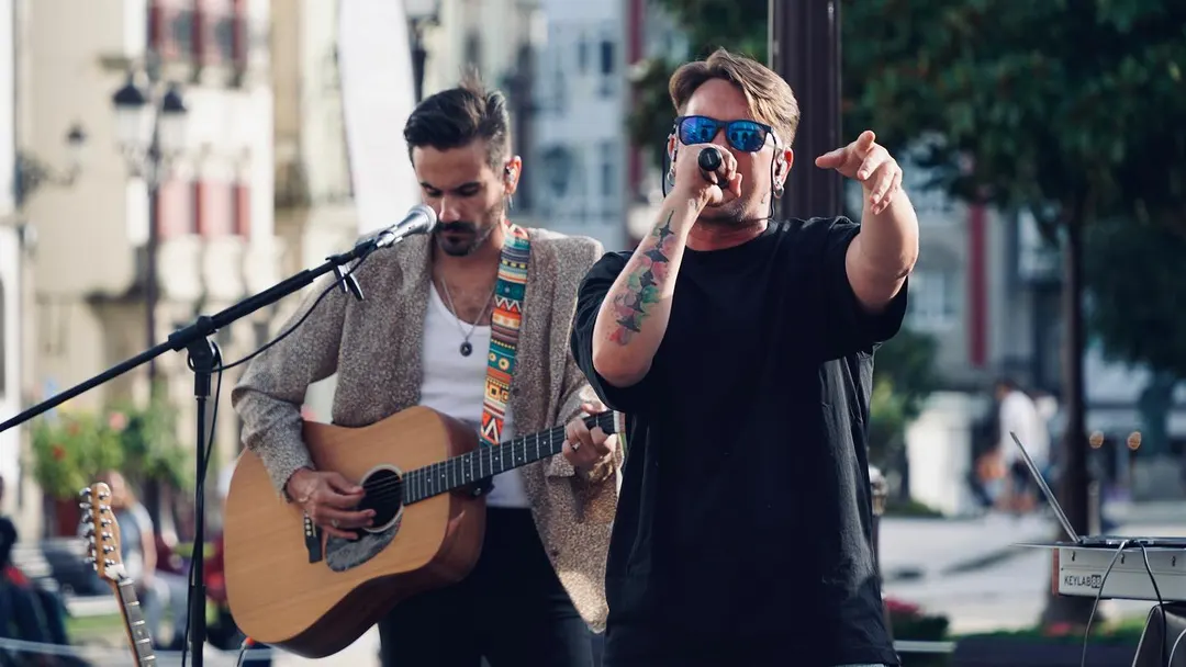 Cedecé cantando en la calle con gafas de sol azules y el brazo señalando al frente, con Rober Carcos a la guitarra detrás.