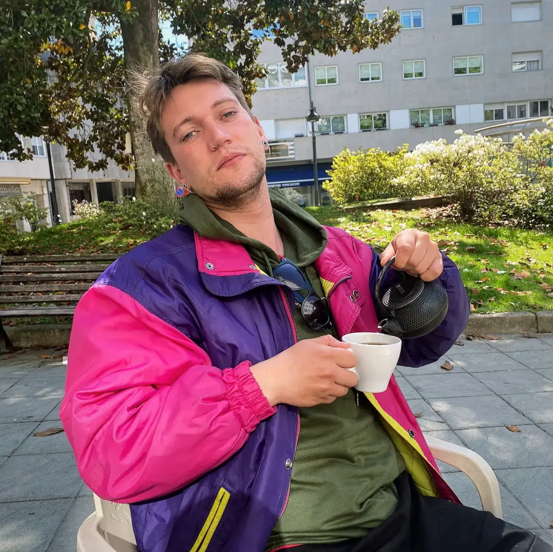 Cedecé sentado en un parque con una cazadora rosa y violeta, sirviéndose un café de una tetera negra.