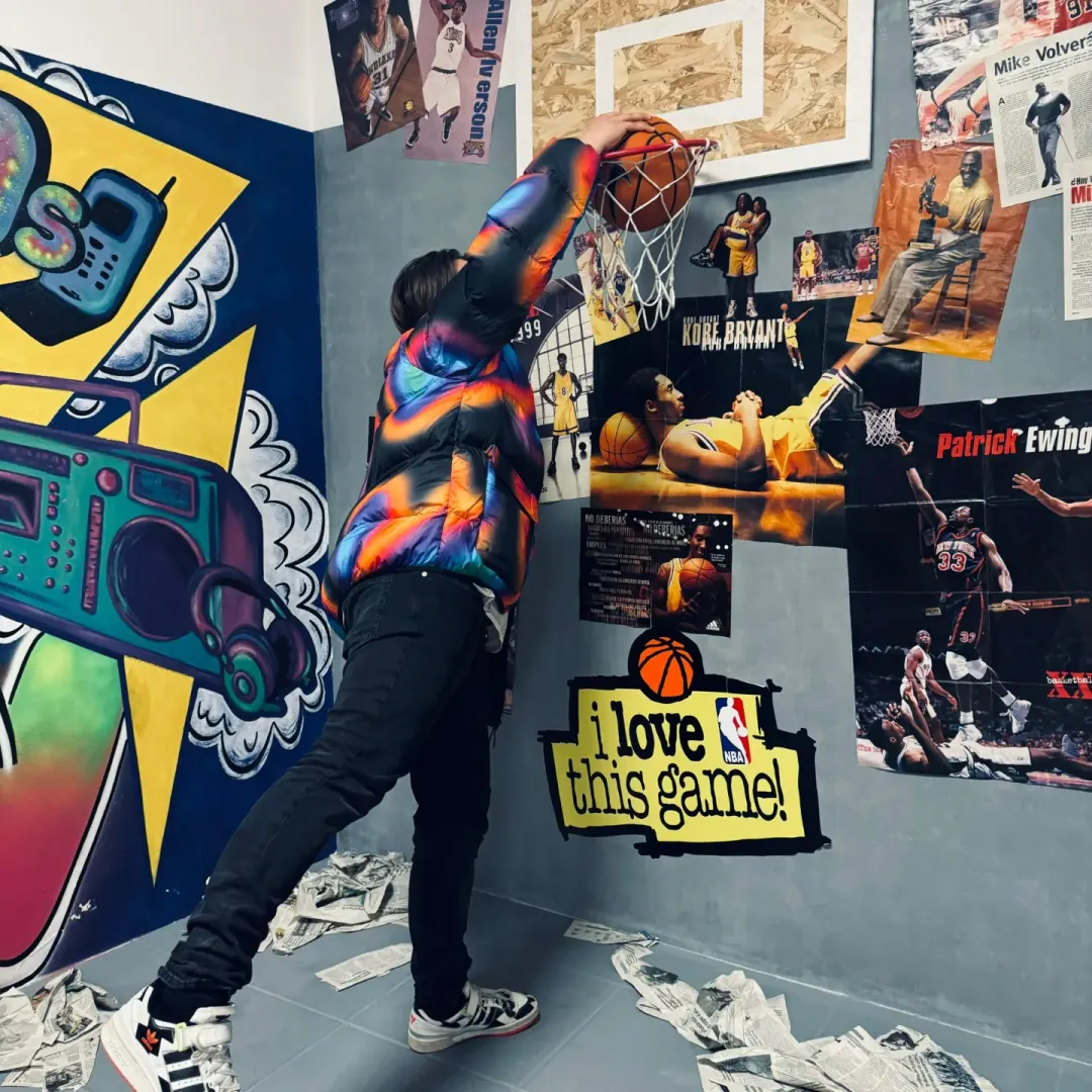 Cedecé machacando un balón en una canasta, junto a un mural de un radiocasete y una pared de carteles de baloncesto.